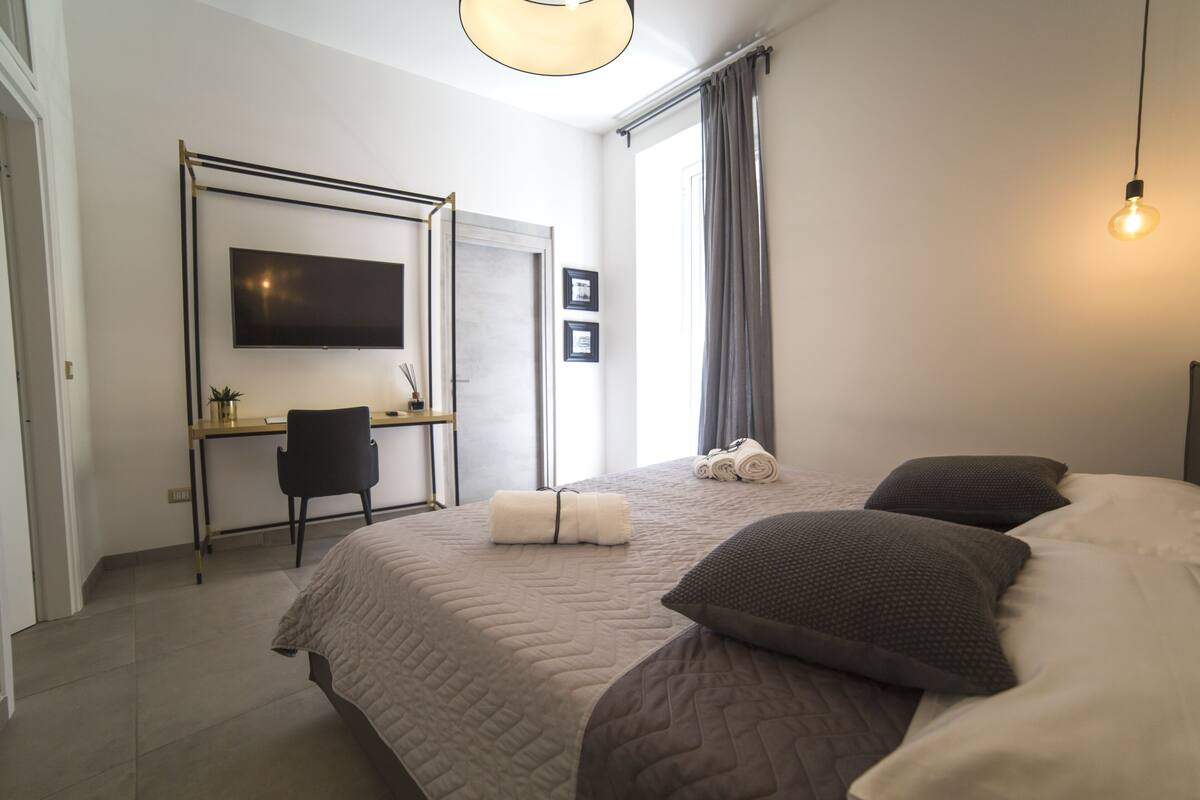 A bedroom with a bed, a desk, a chair, a TV, and a window with curtains.
