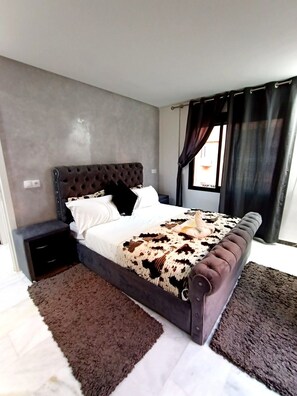 A modern bedroom with a tufted headboard, a large bed with a patterned blanket, a nightstand, and a window with curtains.