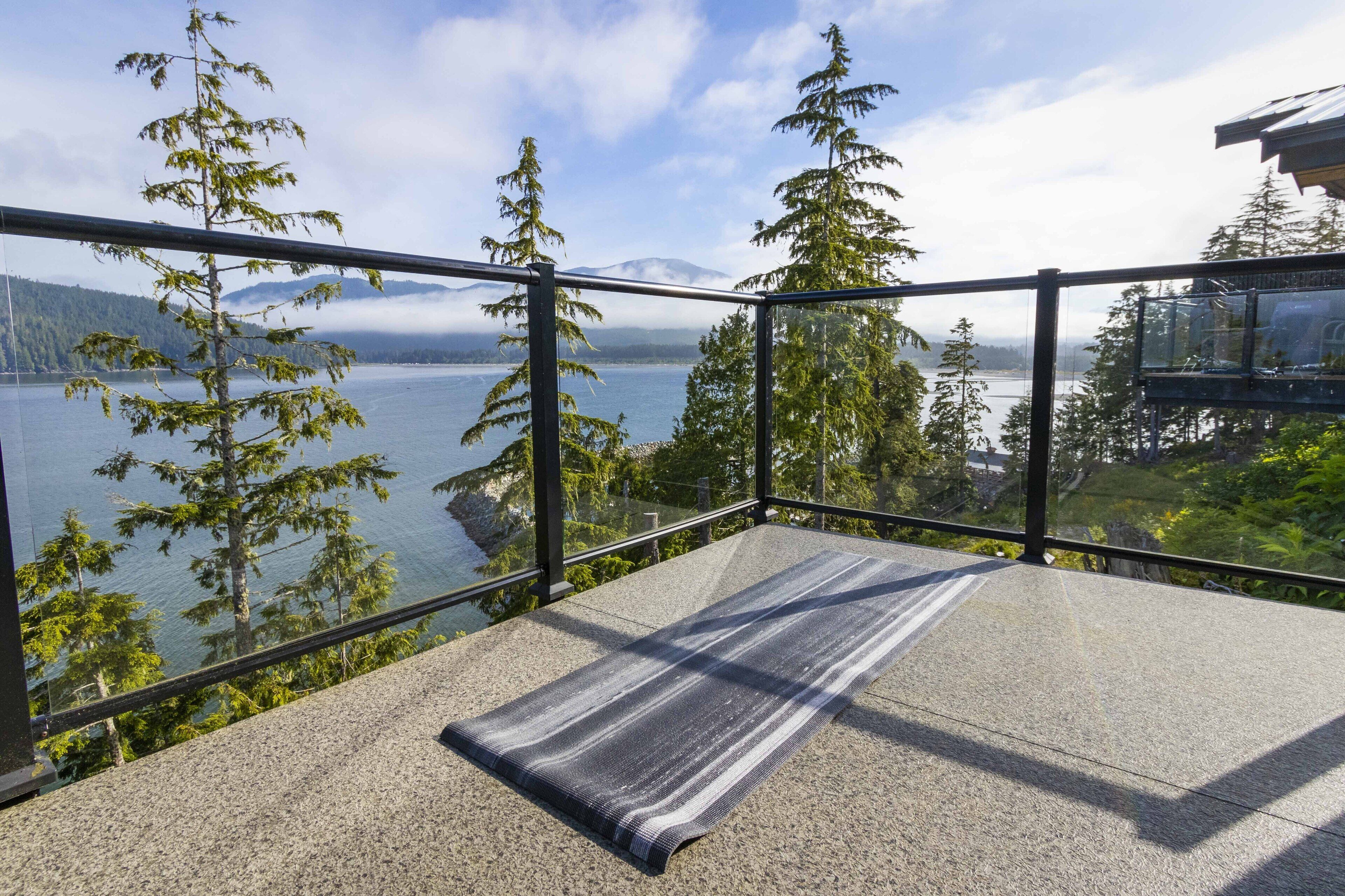 A balcony with a view of a lake and mountains, featuring a glass railing and a mat.