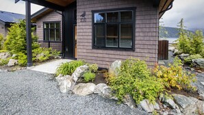 Une maison moderne dotée d’un porche abrité, portant le numéro 230, entourée d’une allée en gravier et d’un jardin aménagé avec des rochers et des arbustes.
