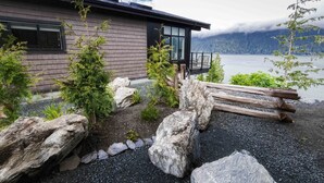 Une maison moderne dotée d’une terrasse donnant sur un lac, entourée de rochers et de verdure.