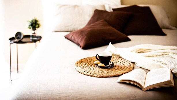 A neatly made bed with pillows, a cup of coffee, and an open book on a woven placemat. - Fabrì 171 (Pescara)