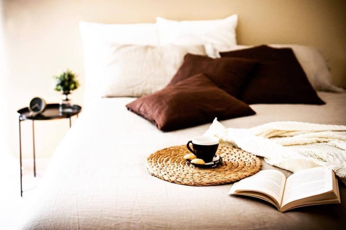 A neatly made bed with pillows, a cup of coffee, and an open book on a woven placemat.