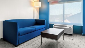 Une pièce avec un canapé bleu, une petite table, un lampadaire et une grande fenêtre donnant sur des immeubles.