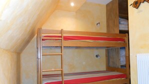 Ein Schlafraum mit Etagenbetten, einer Holzleiter, einem kleinen Tisch mit gemustertem Tischläufer und einem Heizkörper an der Wand.