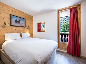 A bedroom with a bed, a wooden wall, a window with red curtains, and a balcony with a railing.