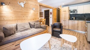A modern living room with a sofa, two white tables, a leather chair, wooden walls, a kitchen with a microwave, and a door leading to another room. - Renovated apartment in the heart of Val-d'Isère (Val-d'Isère)