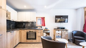 A modern kitchen with wooden cabinets, a built-in oven, and a coffee maker. There is a dining area with a table and chairs, a television mounted on the wall, and a red curtain. - Renovated apartment in the heart of Val-d'Isère (Val-d'Isère)
