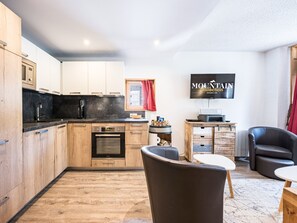 A modern kitchen with wooden cabinets, a built-in oven, and a coffee maker. There is a dining area with a table and chairs, a television mounted on the wall, and a red curtain.