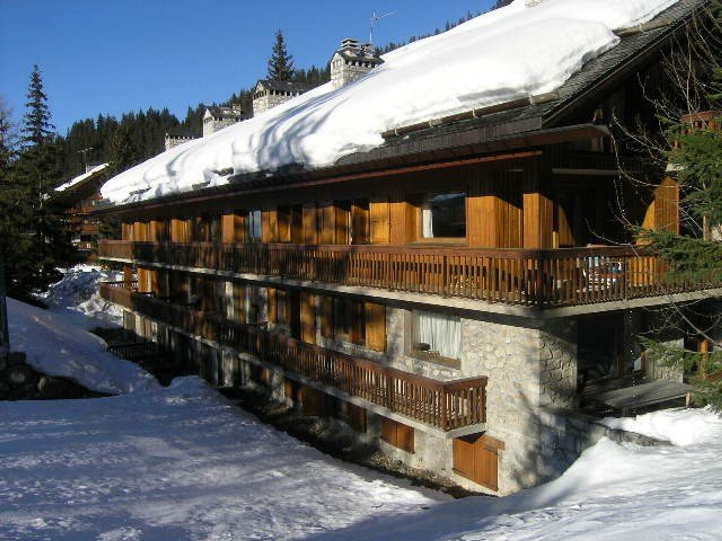 A wooden chalet with a balcony, surrounded by snow-covered ground and trees.