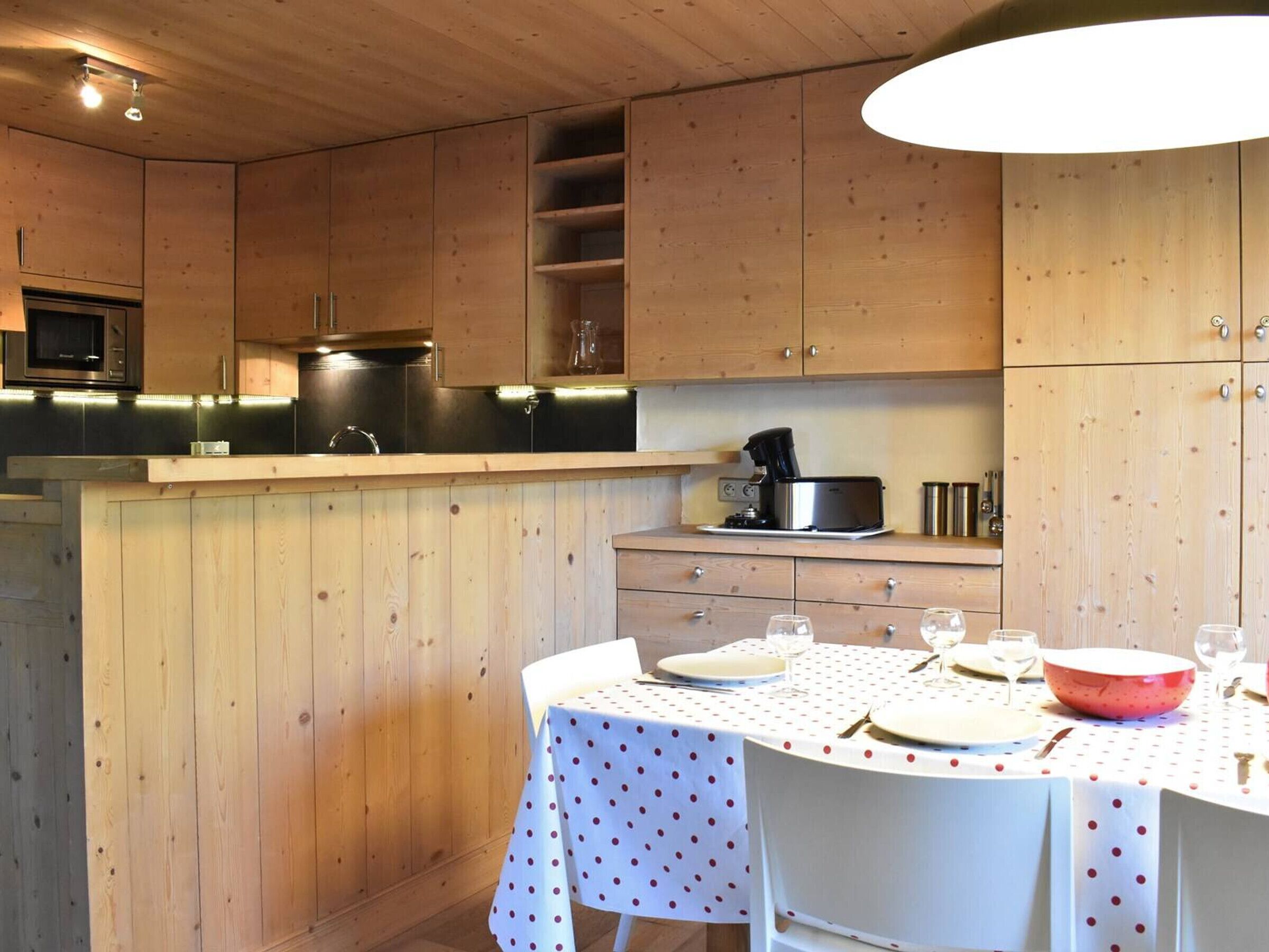 Una cocina con armarios de madera, microondas, cafetera y una mesa de comedor para dos con mantel de lunares.