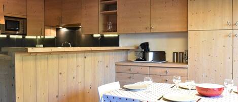 Una cocina con armarios de madera, microondas, cafetera y una mesa de comedor para dos con mantel de lunares.