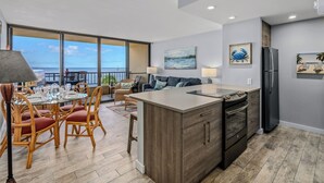 A modern kitchen with a large island, wooden chairs, and a dining table with chairs. There is a view of the ocean through large windows. - Sugar Beach PH31 Front Row Amazing Oceanviews in Highly Upgraded Unit (Kihei)