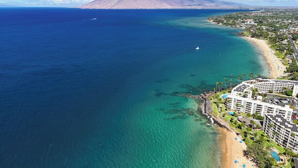 Luchtbeeld van een kustgebied met een strand, een hotel met meerdere verdiepingen en een berg op de achtergrond.