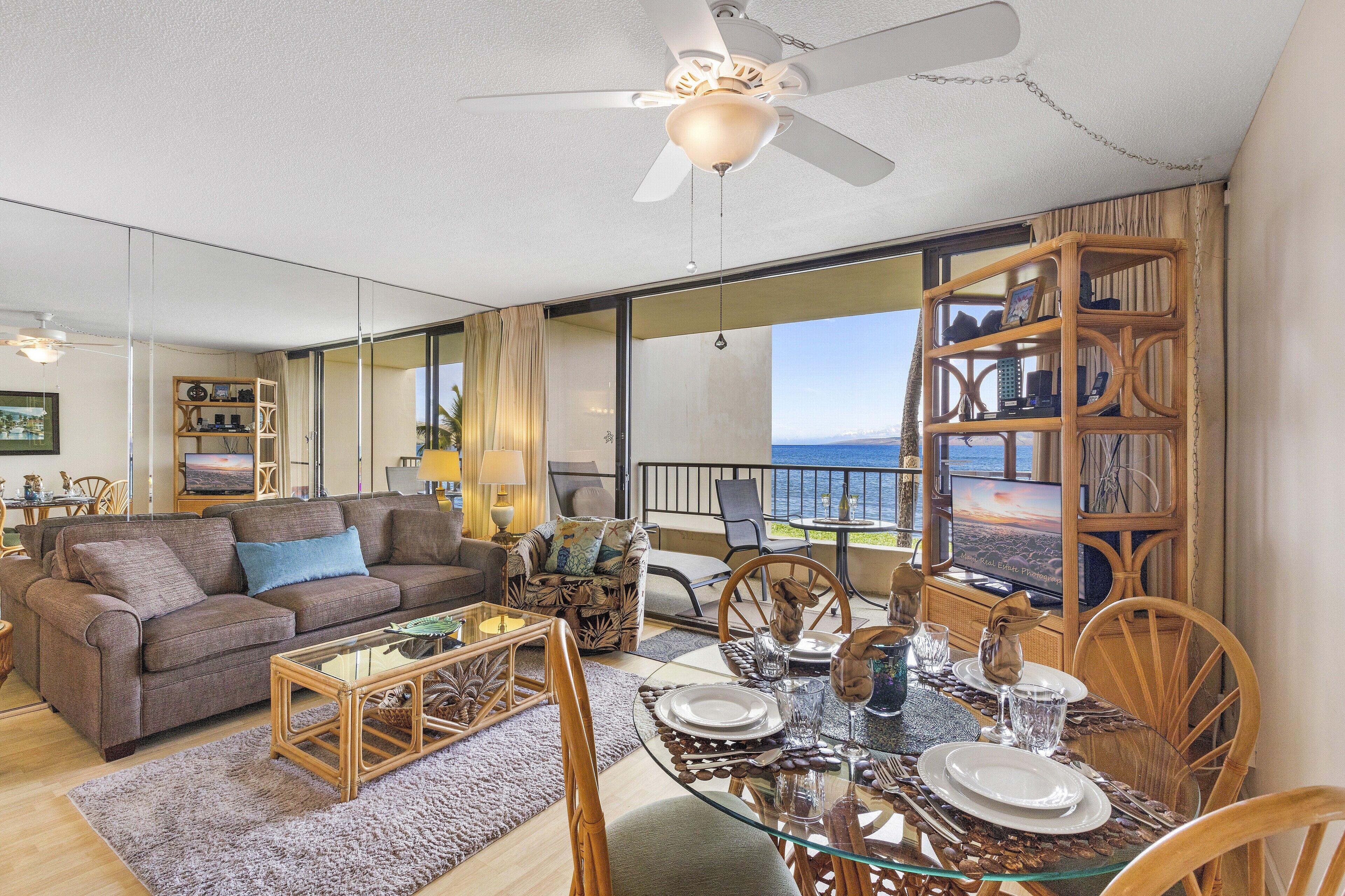 A spacious living area with a large window overlooking the ocean, a ceiling fan, and a dining table set for four.