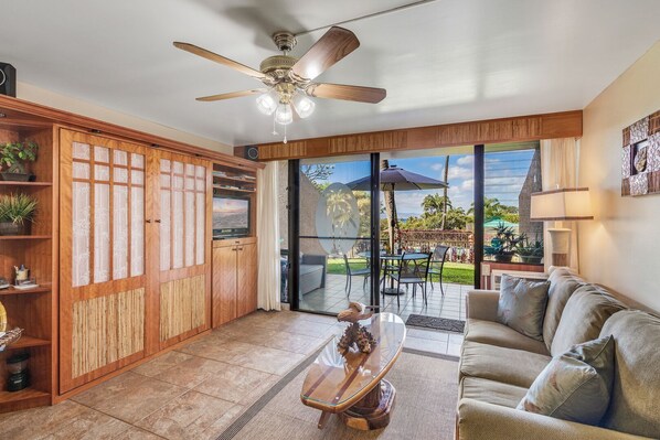 A spacious living area with a ceiling fan, a glass coffee table, a sofa, and a sliding glass door leading to an outdoor patio.