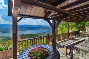 Una pérgola de madera con un banco de piedra y una mesa, con vistas a un lago y montañas panorámicos.
