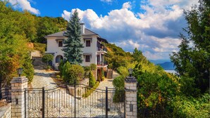 A two-story house with a stone pathway leading to a gated entrance, surrounded by lush greenery and a view of the sea. - Infinity Villa Ioannina (Ioannina)