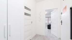 A hallway with white doors, a light wooden floor, and a view into a room with a bed and a desk.