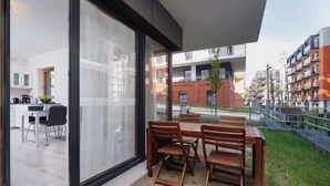 A modern outdoor dining area with a table and chairs, large glass sliding doors, and a view of a cityscape with buildings and a brick structure.