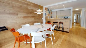 Ein moderner Essbereich mit einem Marmortisch, weißen Stühlen und einem orangefarbenen Sessel. Eine Holzwand und eine Küche mit Barbereich sind sichtbar.