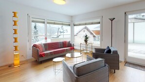 Ein modernes Wohnzimmer mit einer Couch, Sesseln, einem Couchtisch und einer Stehlampe. Große Fenster bieten Blick auf eine verschneite Landschaft.