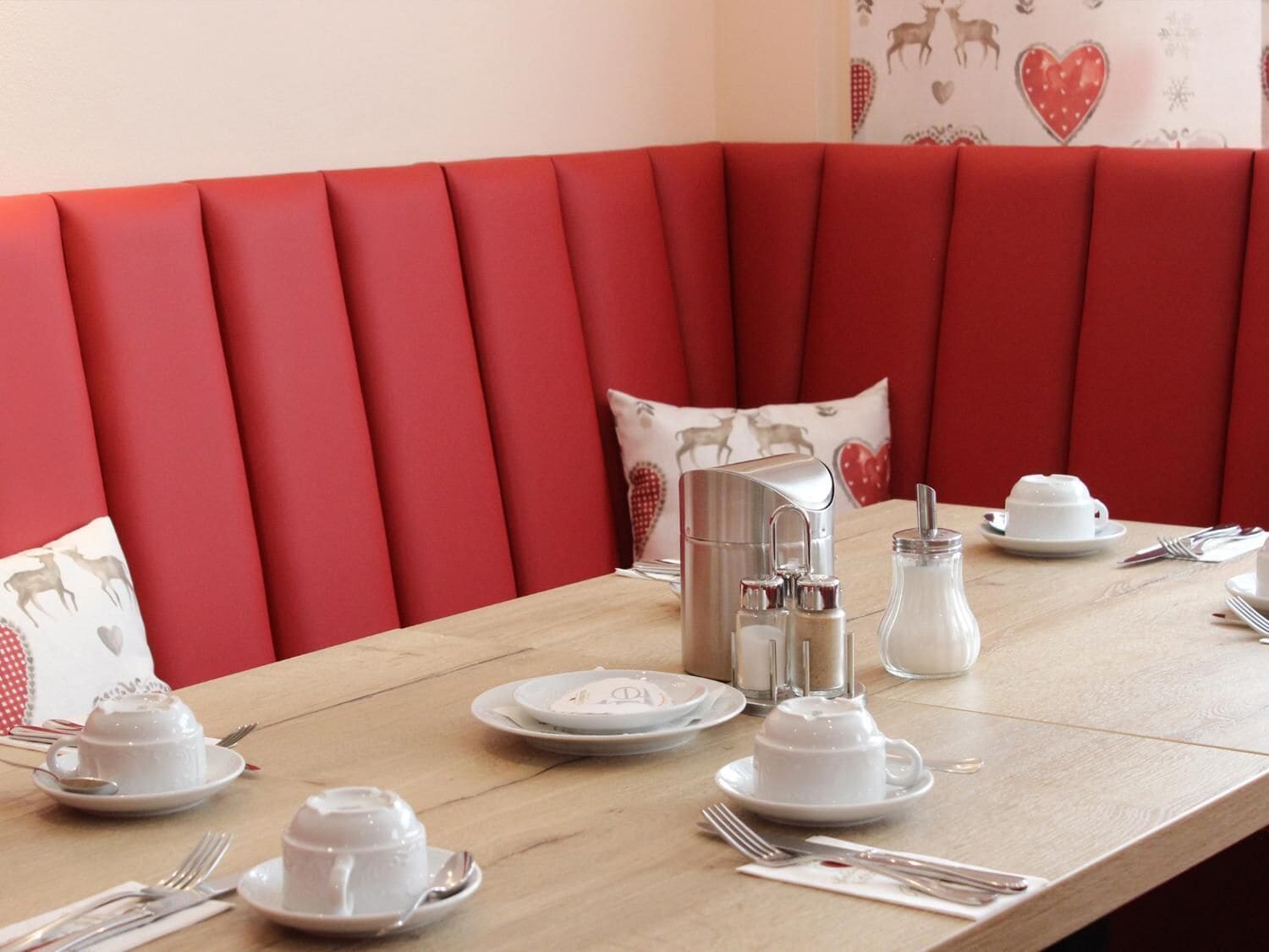 A dining table set with white tableware and a silver pitcher, surrounded by red upholstered seating.