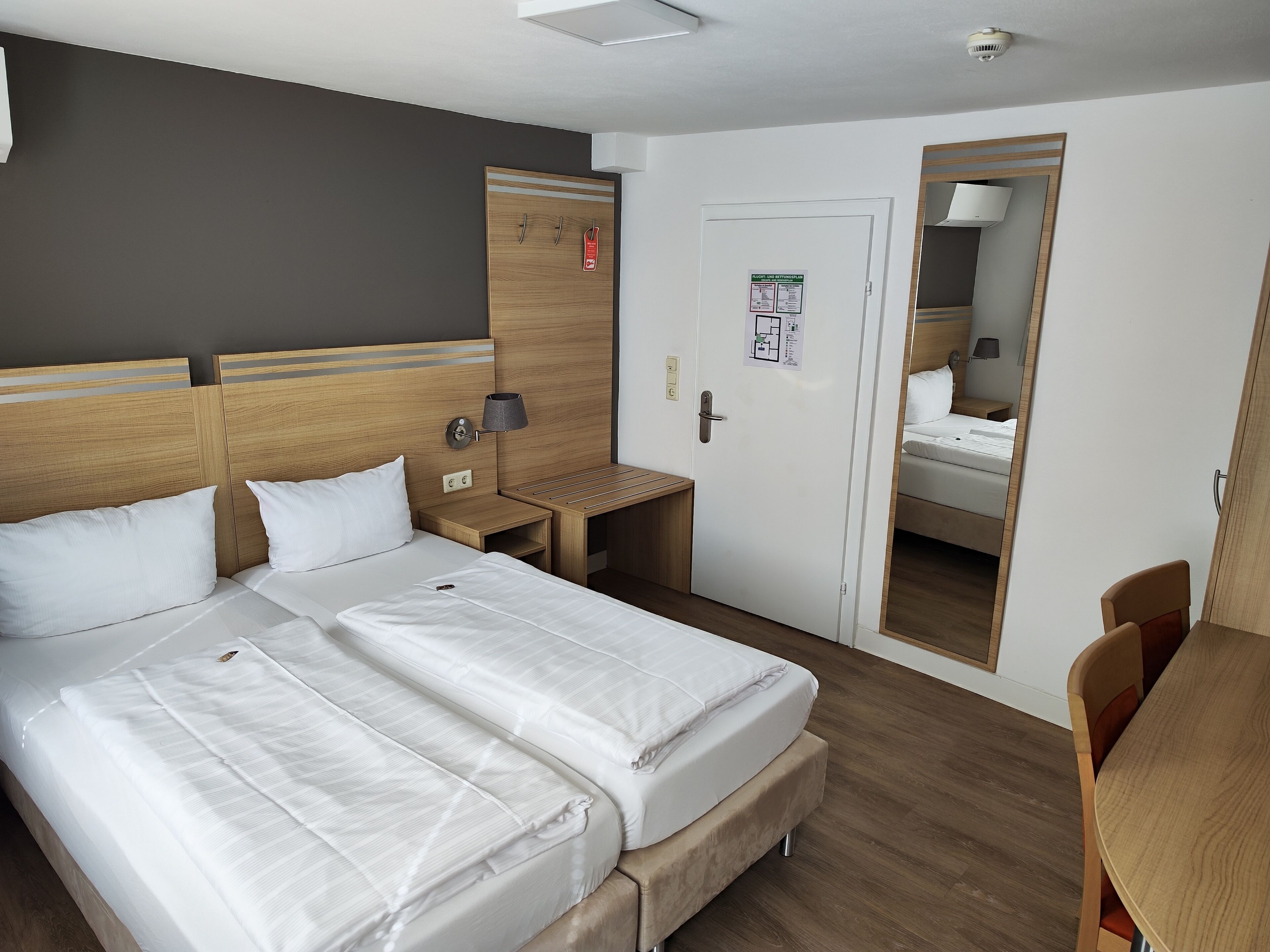 A hotel room with two beds, a desk, a chair, a mirror, and a door with a notice.