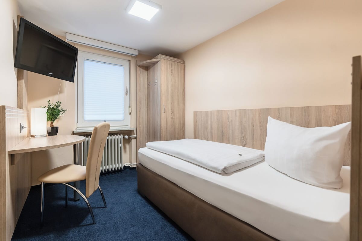Habitación de hotel pequeña con una cama individual, un escritorio con silla, un televisor montado en la pared y una ventana con persianas.