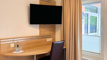 Ein Hotelzimmer mit Flachbildfernseher an der Wand, einem Holztisch mit Stuhl und einem Fenster mit Vorhängen.