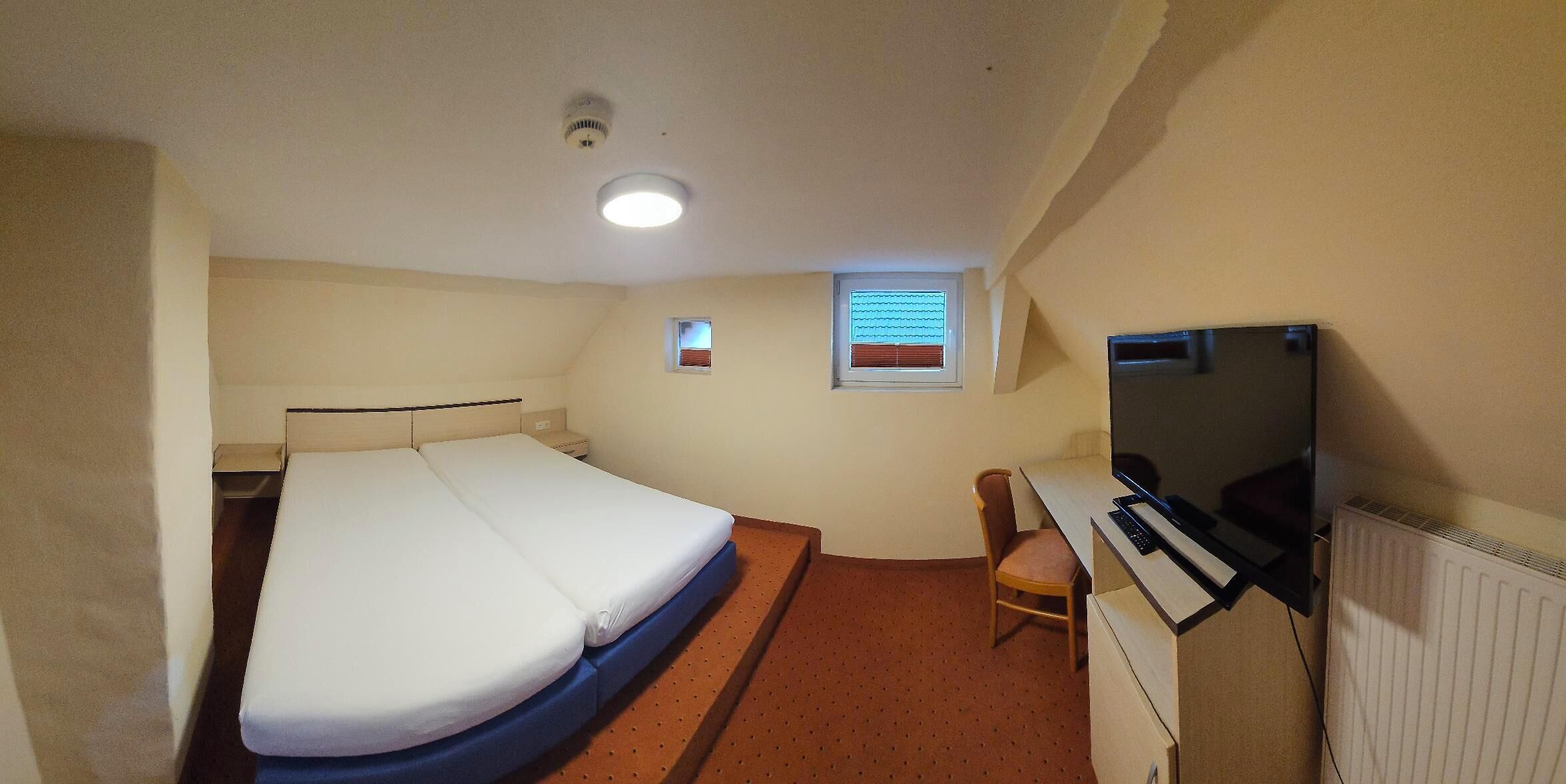 A small, single-bed room with a desk, chair, television, and a window with blinds.