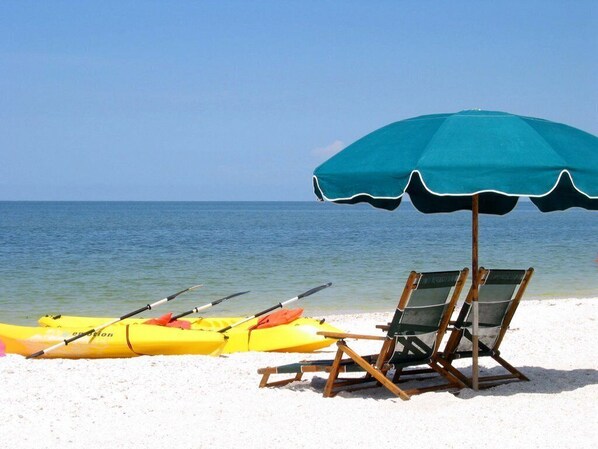 A beach scene with a blue umbrella, two kayaks, and a beach chair.
