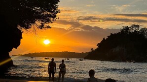 Une vue au coucher du soleil, avec des personnes près de l’eau, une côte rocheuse et la silhouette d’un arbre.