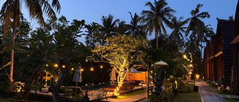 A tropical resort with a paved pathway, palm trees, string lights, and a building with a tiled roof.