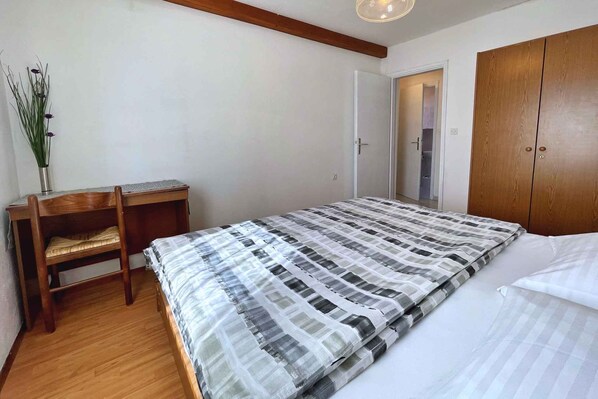 A bedroom with a bed, a wooden wardrobe, a desk with a chair, and a small plant.