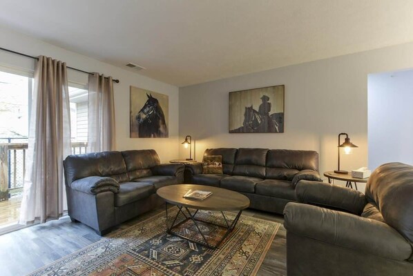 A living room with a large L-shaped sofa, a round coffee table, and two side tables with lamps. There are two wall-mounted paintings and a view of the outdoors through a window with curtains. - Western Comfort (Branson)