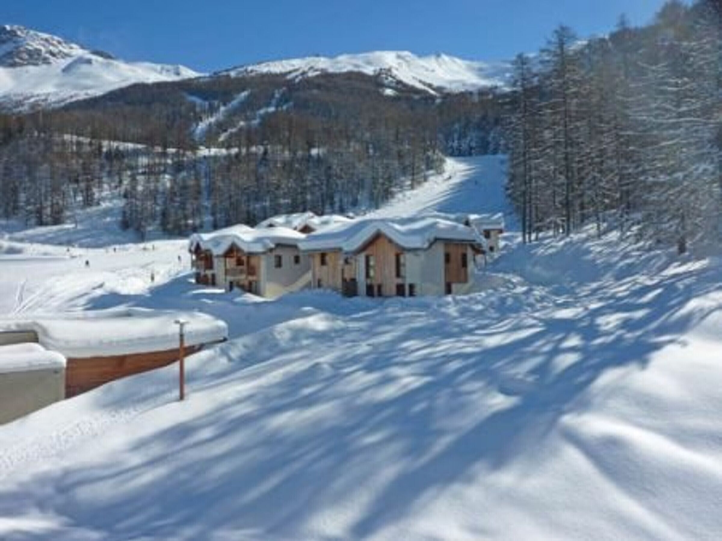 A snow-covered lodge with multiple cabins, a ski slope in the background, and a forested mountain range.