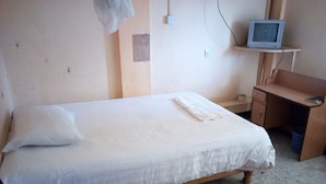 A single-bed room with a wooden desk, a television on a shelf, and a patterned rug. - Olympia Hotel (Nyahururu)