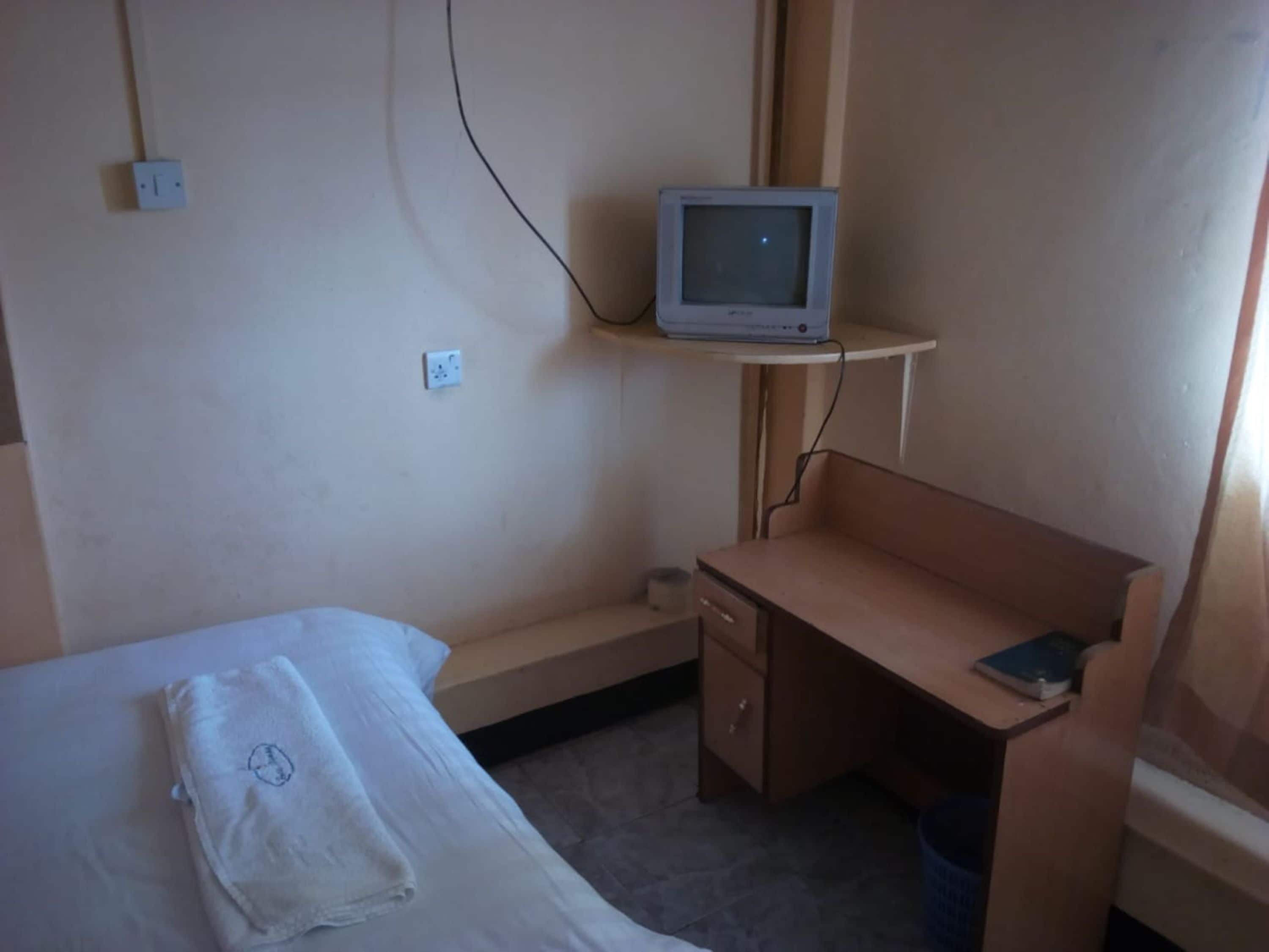Een kleine, eenvoudige kamer met een bed, een bureau en een televisie op een plank.