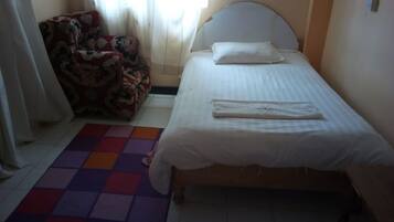A small, neatly made bed with a white headboard and a folded white towel on it. There is a colorful rug on the floor and a floral-patterned chair to the left.