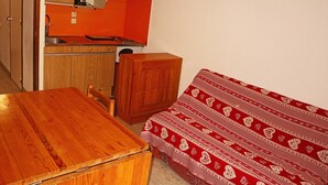 A compact kitchen with wooden cabinets, a sink, and a small countertop. There is a wooden table and a single bed with a patterned blanket. - Studio 6 pers avec mezzanine, accès pistes, proche commerces, balcon Est (Les Orres)