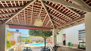 Un espace extérieur aménagé en terrasse, avec des tables et des chaises en bois, un sol carrelé et une vue sur une piscine et la verdure.