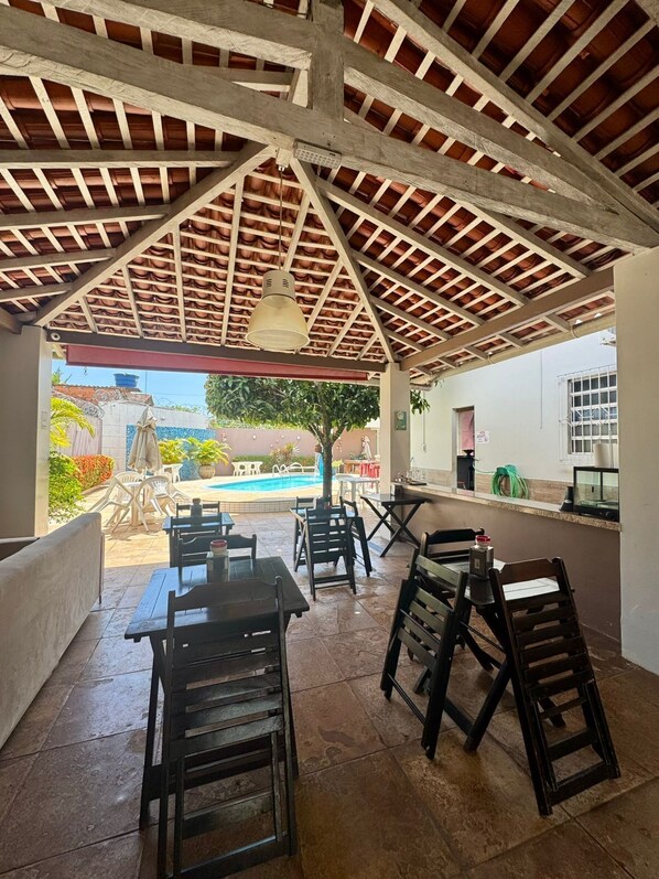 An outdoor patio area with wooden tables and chairs, a tiled floor, and a view of a swimming pool and greenery. - Porto Plaza Pousada (Barcarena)