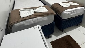 Une petite chambre, de taille modeste, avec deux lits simples, un climatiseur blanc fixé au mur et une petite table surmontée d’un tapis marron.
