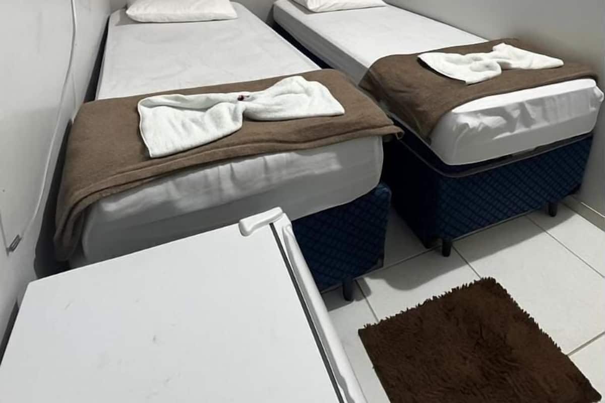 Une petite chambre, de taille modeste, avec deux lits simples, un climatiseur blanc fixé au mur et une petite table surmontée d’un tapis marron.