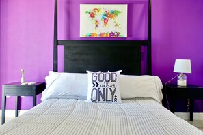 A bed with striped bedding, a white pillow with "GOOD vibes ONLY" text, a black headboard, and a purple wall with a world map artwork.