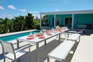 An outdoor dining area with white tables and chairs, a pool, and a turquoise building in the background.
