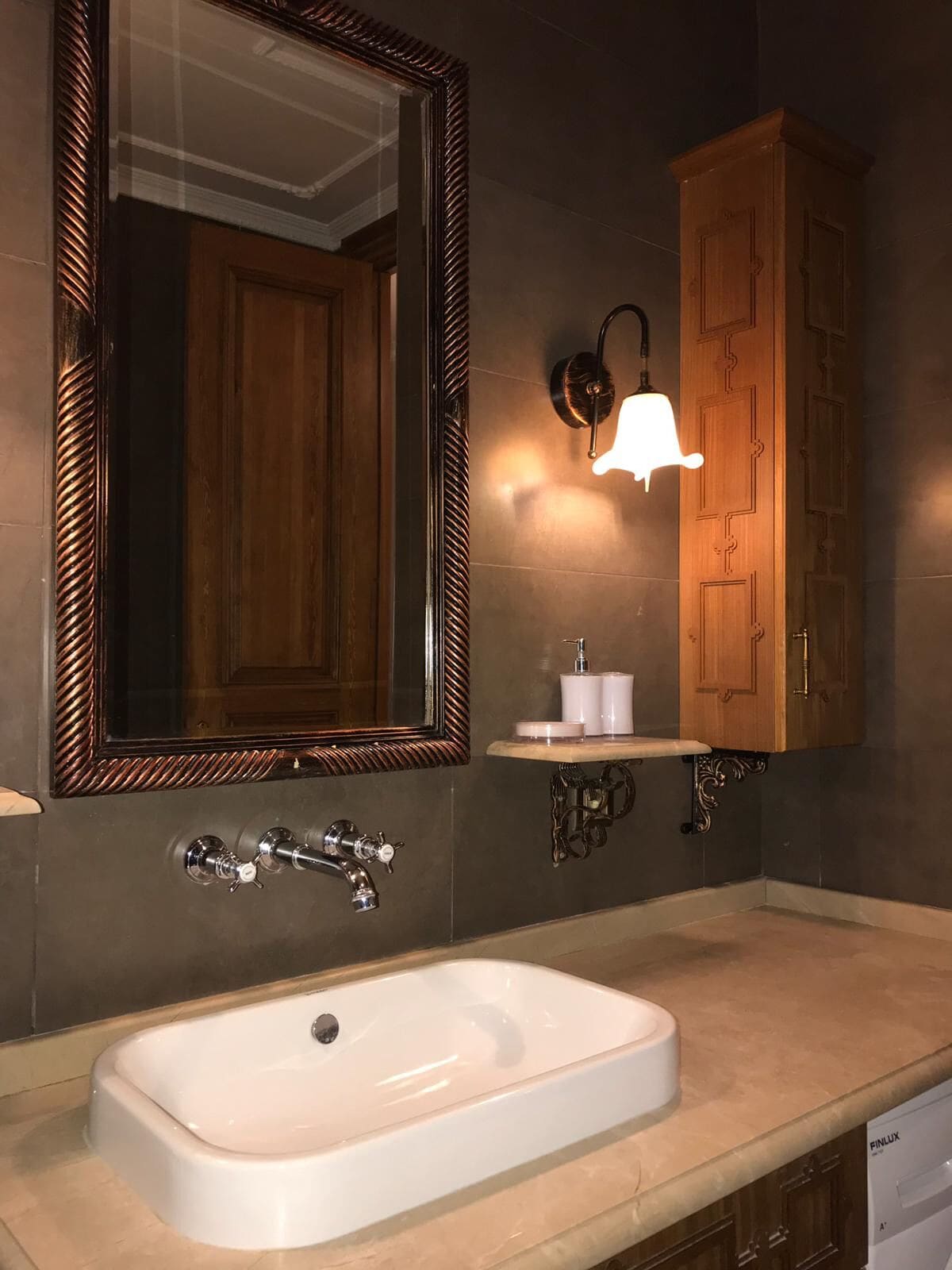 Un baño con un espejo grande, un lavamanos blanco, un gabinete de madera y una lámpara empotrada en la pared.