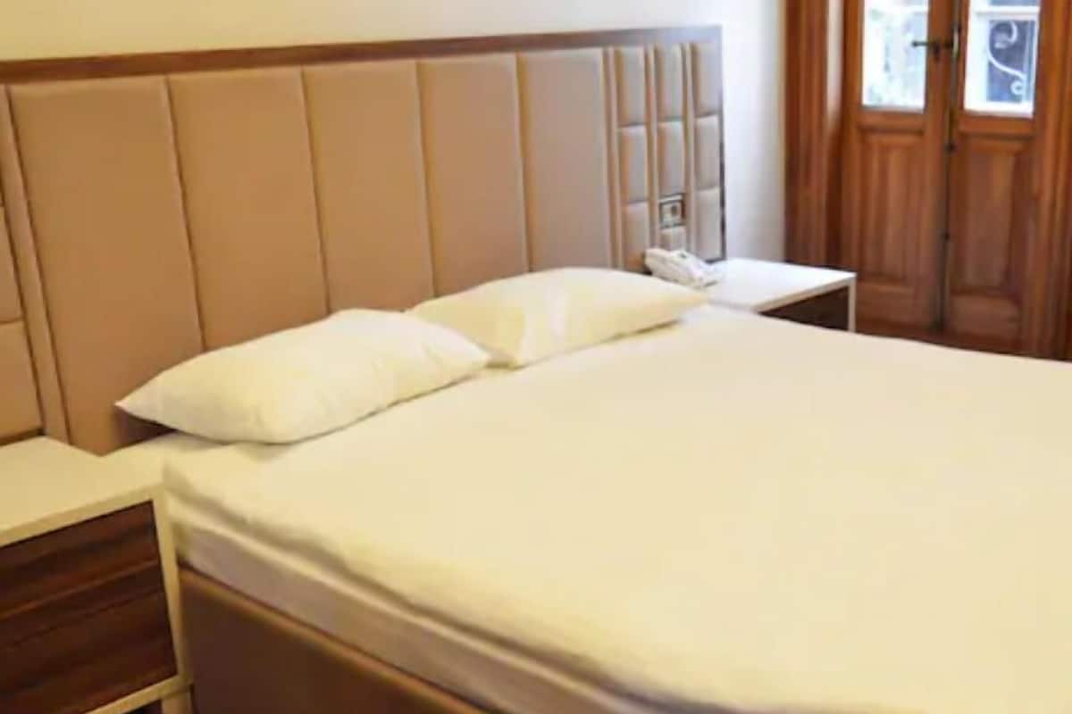 A neatly made bed with a wooden headboard, a bedside table with a phone, and a window with curtains.