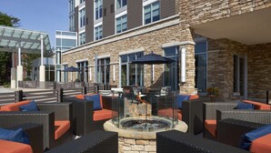 Een terras met rieten meubels en oranje kussens, een vuurkorf, en een stenen gebouw met grote ramen.